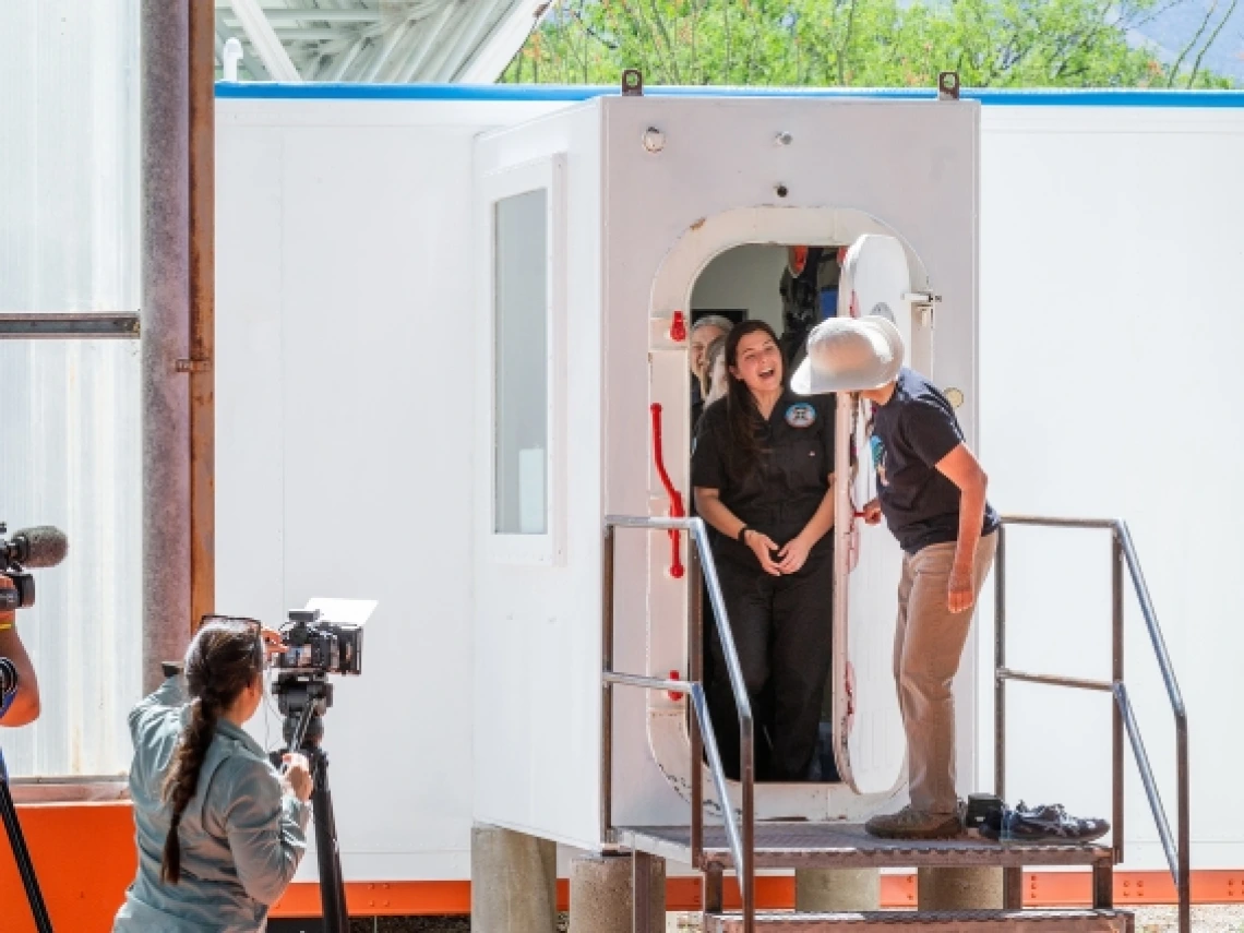 A person emerges from a hatch and is greeted by another standing outside