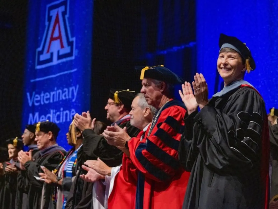 A group of people wearing graduation gowns stand in line on stage and clap in front of a background. 