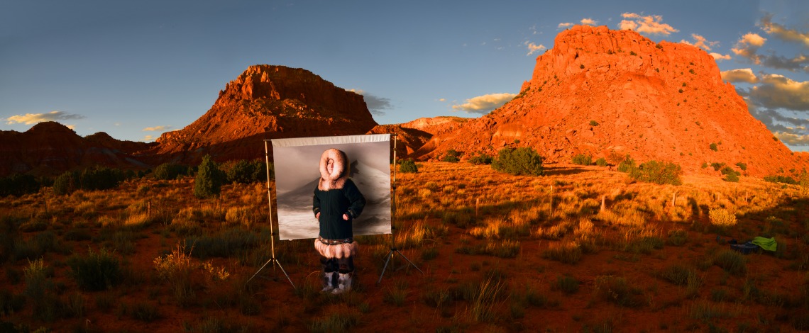 A photograph of poet Joan Kane, in front of a backdrop and a red mountain range at sunset.