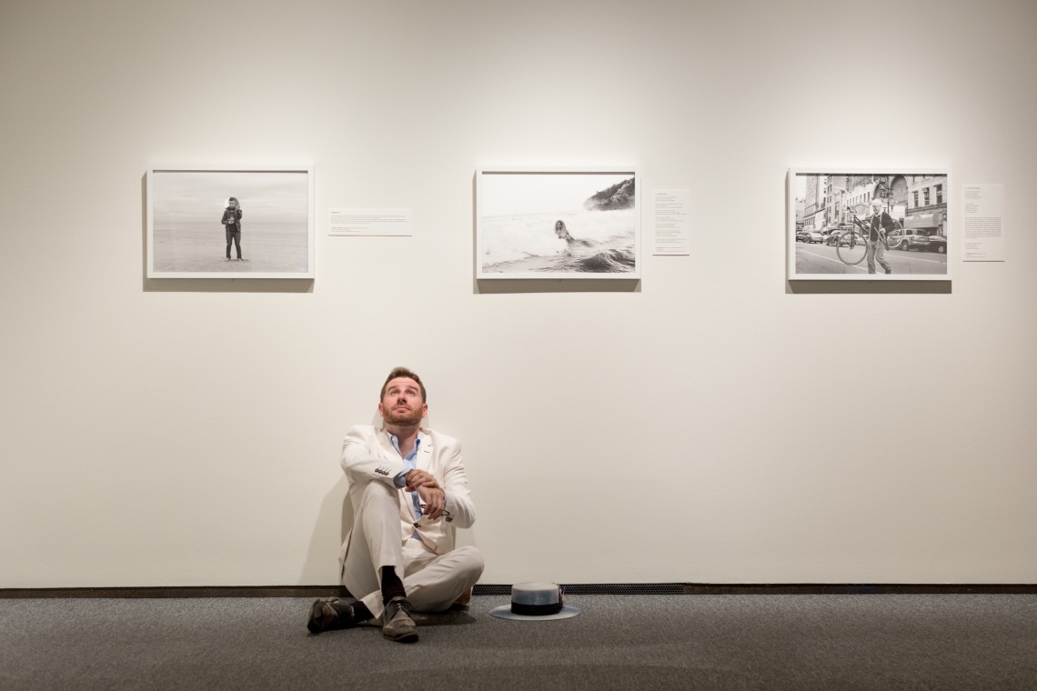 A photograph of Van Sise, sitting on the floor, looking up at the sky, surrounded by his framed photographs