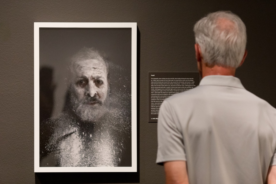 A photograph of a man's back turned, looking at a frame photograph of a man's face covered in dust