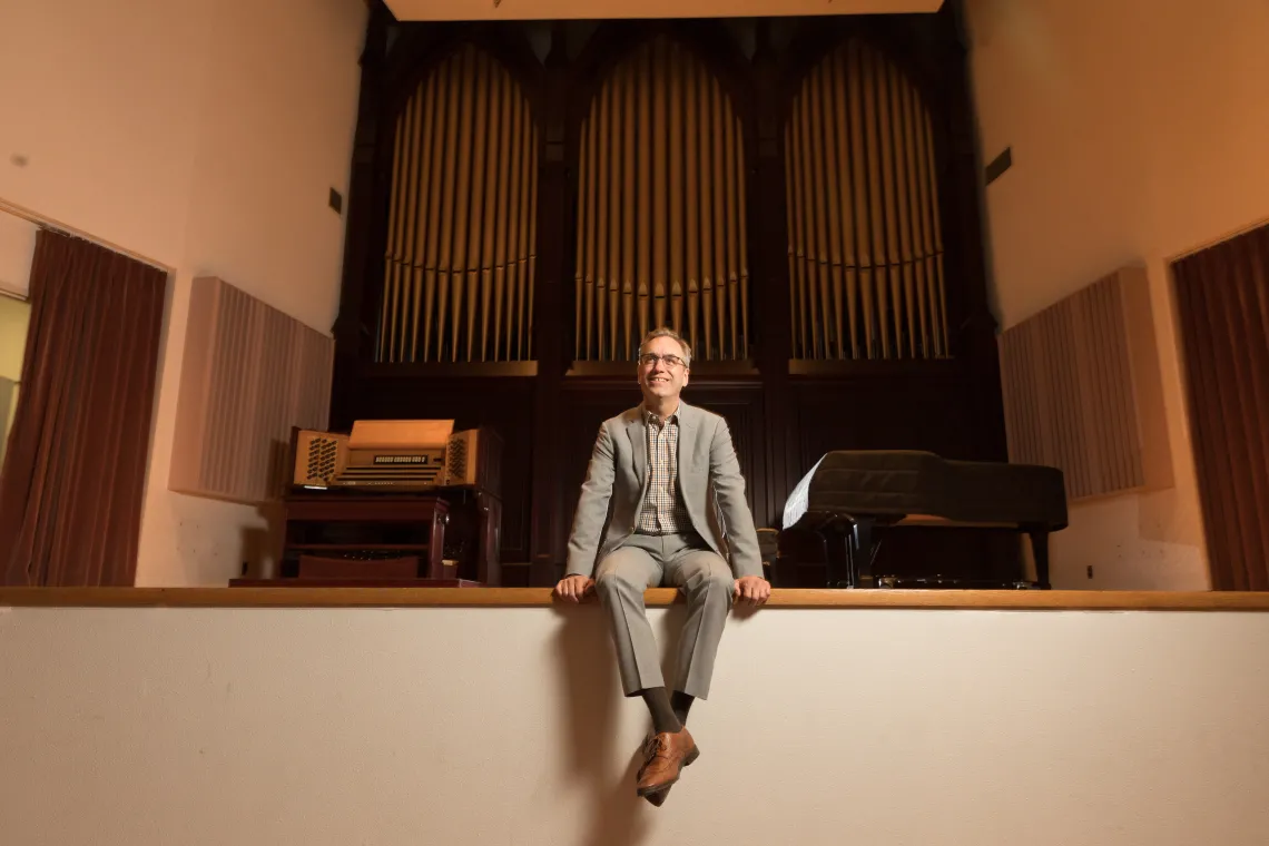 A photograph of Andrew "Andy" Schulz sitting at the edge of the stage, smiling, with his legs crossed.