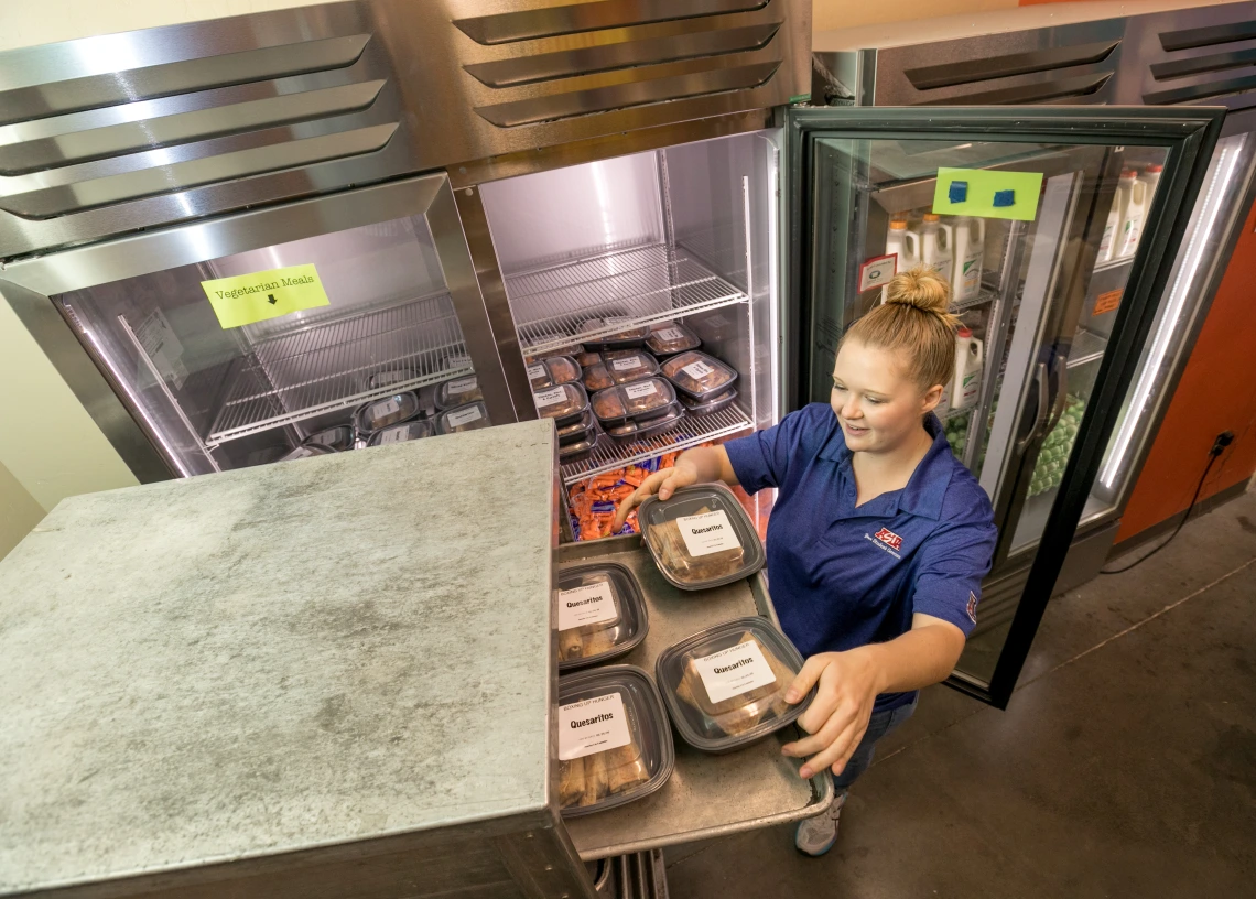 A photograph of a student employee loading ready-to-go meals onto a tray