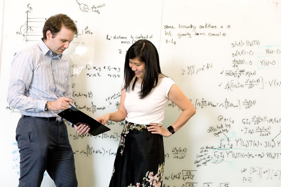 A photograph of Euan McLeod and Judith Su pointing to a clipboard and talking in front of a whiteboard