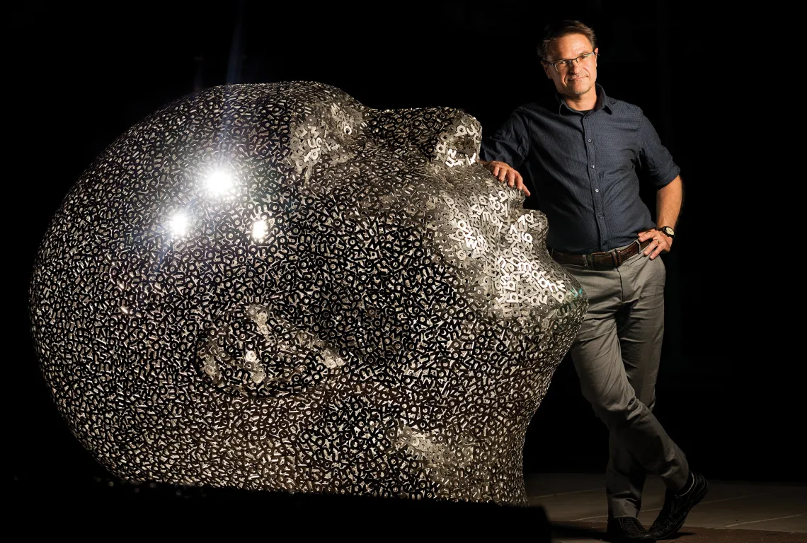 A photograph of Stephen Cowen standing near a human-sized sculpture of a head