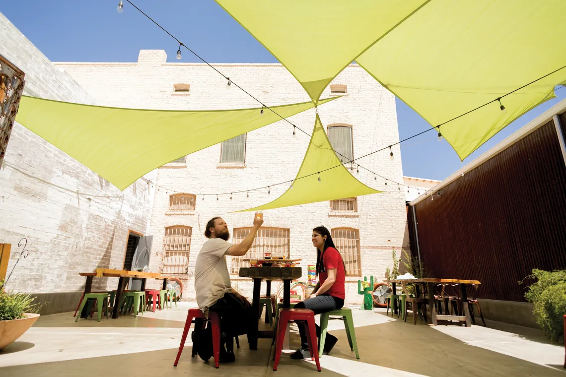 A photograph of two individuals sitting at a table under a shaded area