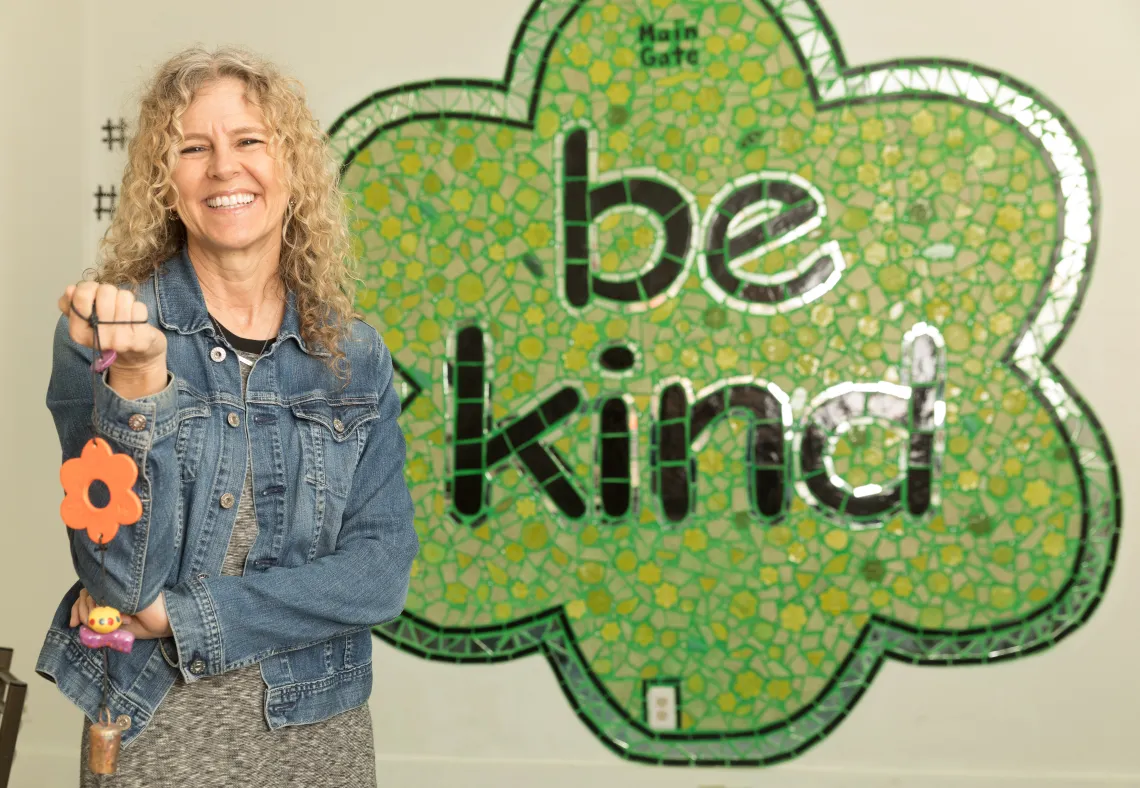 A photograph of Jeannette Maré holding a Ben's Bell in front of a "Be Kind" sign