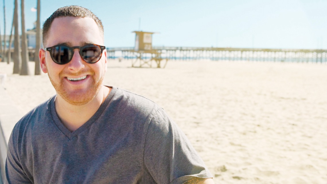 A photograph of Joey Katzen, smiling, wearing sunglasses, on a beach boardwalk.