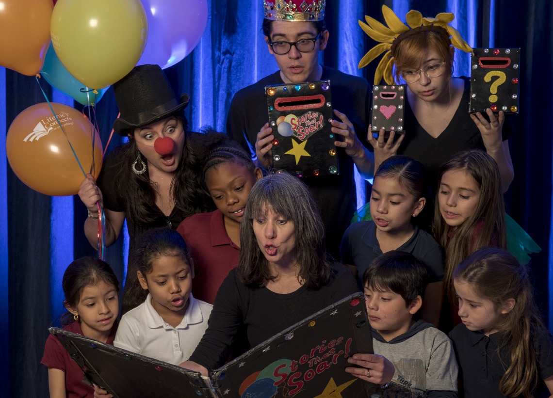 A photograph of Sharon O'Brien reading from a large book. Animated actors wearing silly hats, clown noses and balloons surround her. Children are also gathered around to read.