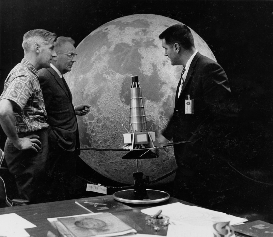 Ewen Whitaker, Gerard Kuiper and Ray Heacock, talking and intently gazing at a model of a Ranger spacecraft.