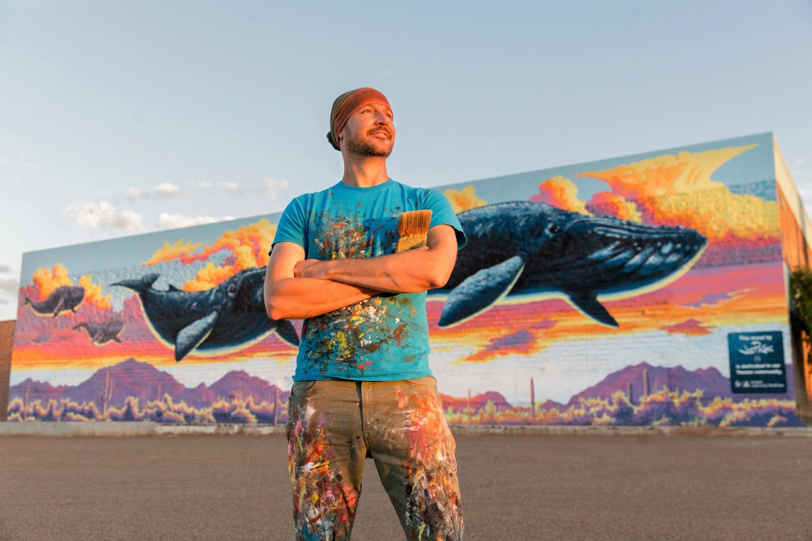 A photograph of Joe Pagac standing and smiling, with a paint brush in his hand and his pants covered in paint. Behind him is a mural of whales swimming in the Sonoran sunset sky.