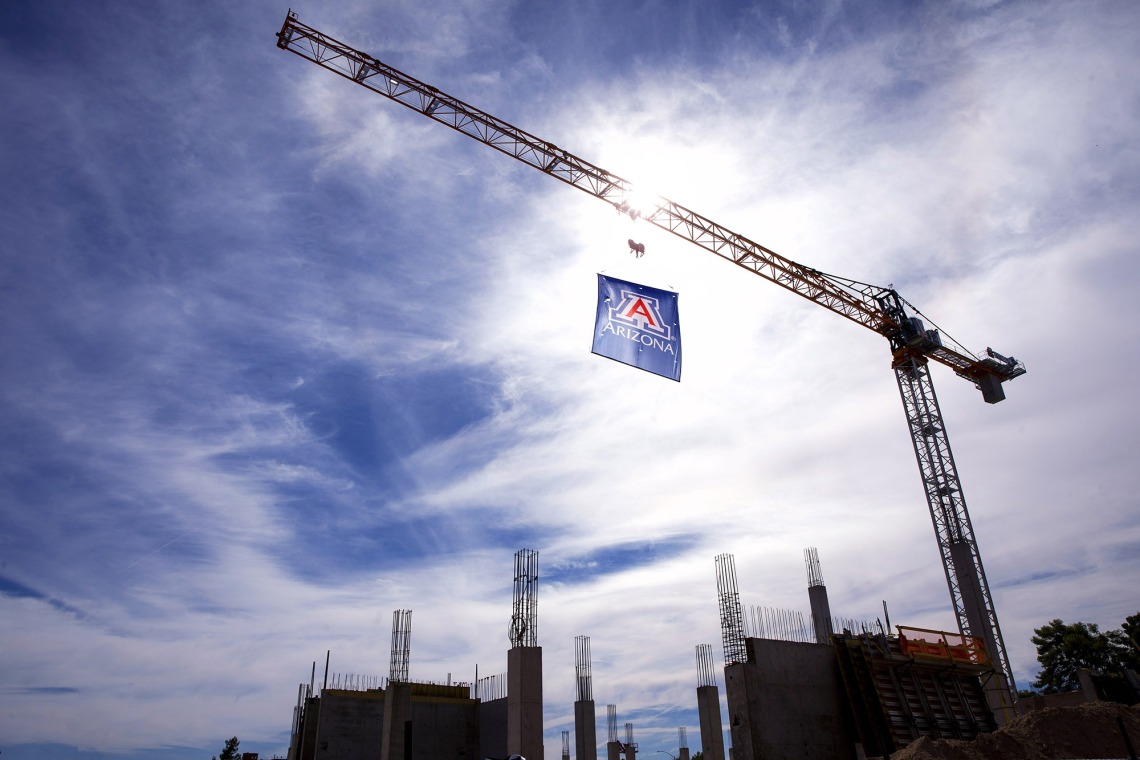 A photograph of a crane holding the University of Arizona Block 'A' Flag against a cloudy backdrop.