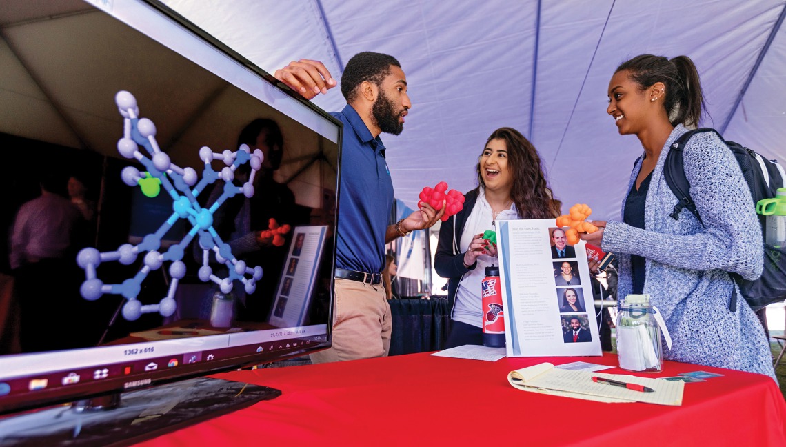 A photograph of three students standing, chatting and staring at a screen at the 2018 McGuire Innovation Expo