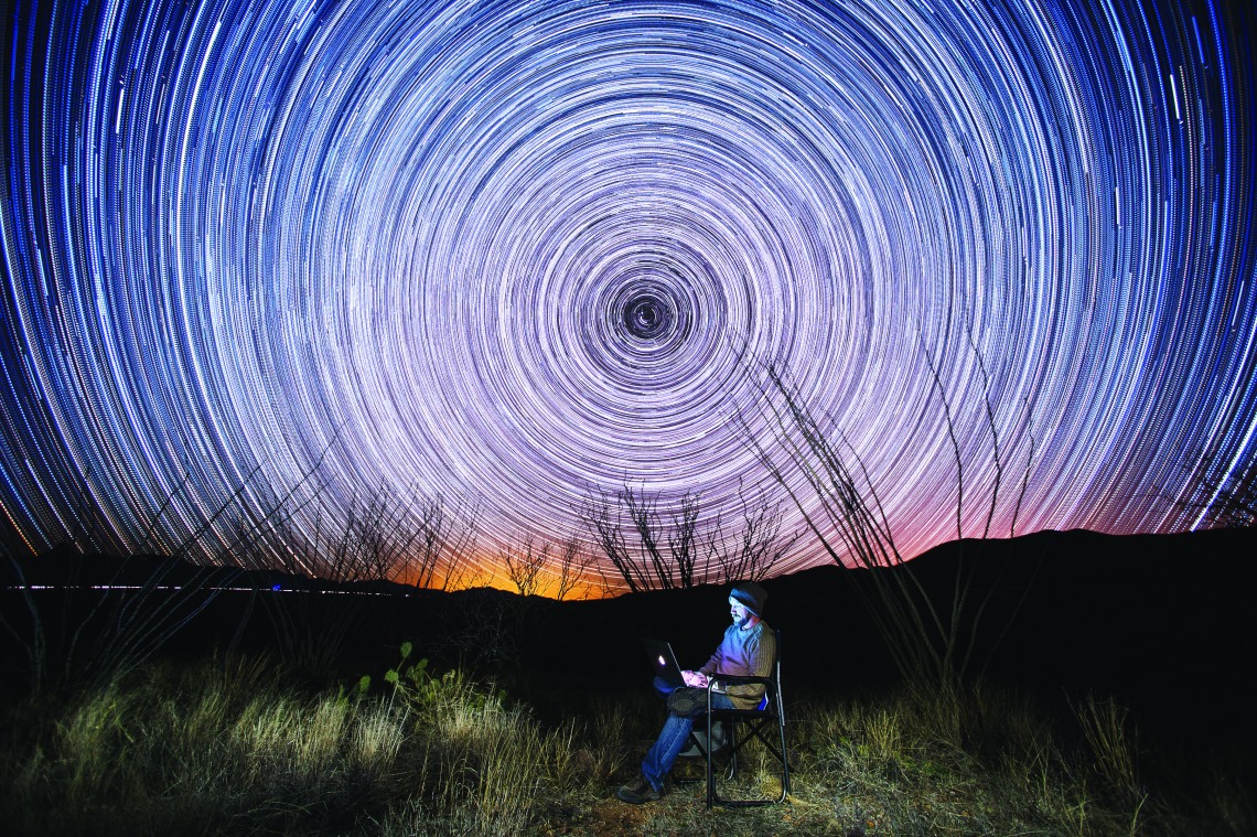 A photograph of a student, sitting with a laptop underneath the Star Trails 