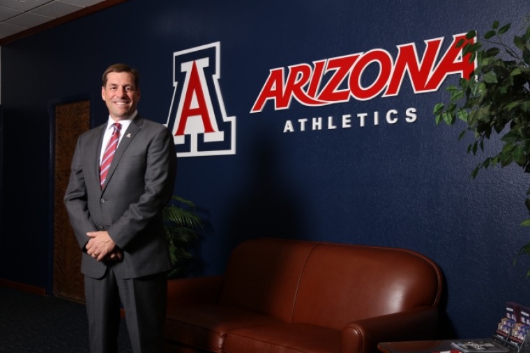 A photograph of Dave Heeke standing in front of an Arizona Athletics wall mural