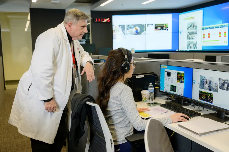 A photograph of a man wearing a white lab coat looking at a computer alongside a woman sitting down with headphones 