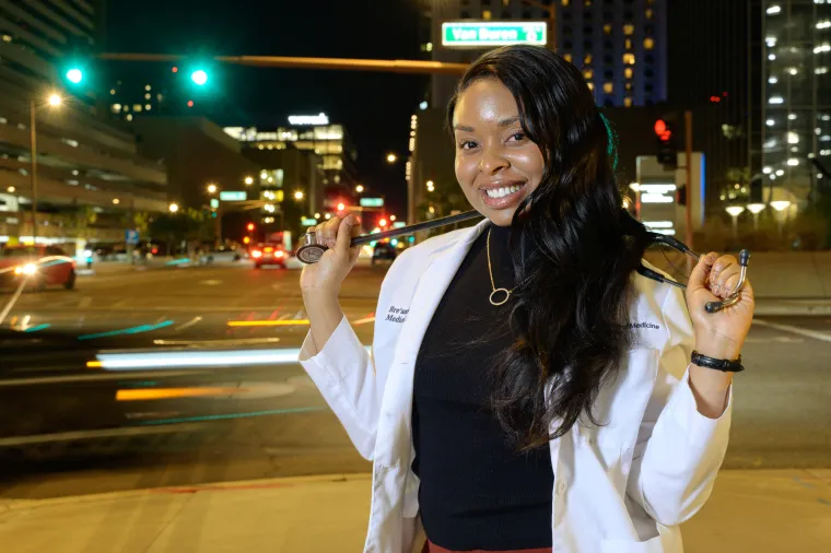 A photograph of a woman standing in a street wearing a white lab coat, holding a stethoscope around her neck. 