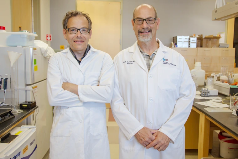 Two men in white lab coats standing in a medical research lab, smiling at the camera.