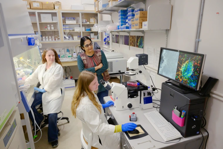 Three women in a research lab, with one seated at a microscope while colorful cellular images display on a monitor.