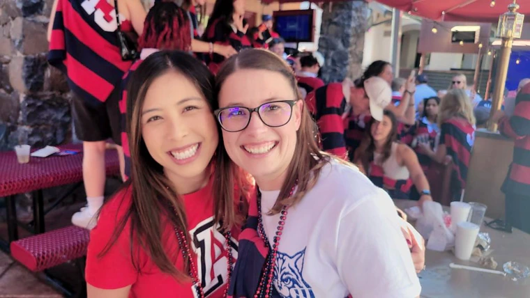 Two people wearing Arizona gear smile and pose together at a lively event filled with people in red and navy striped rugby shirts.