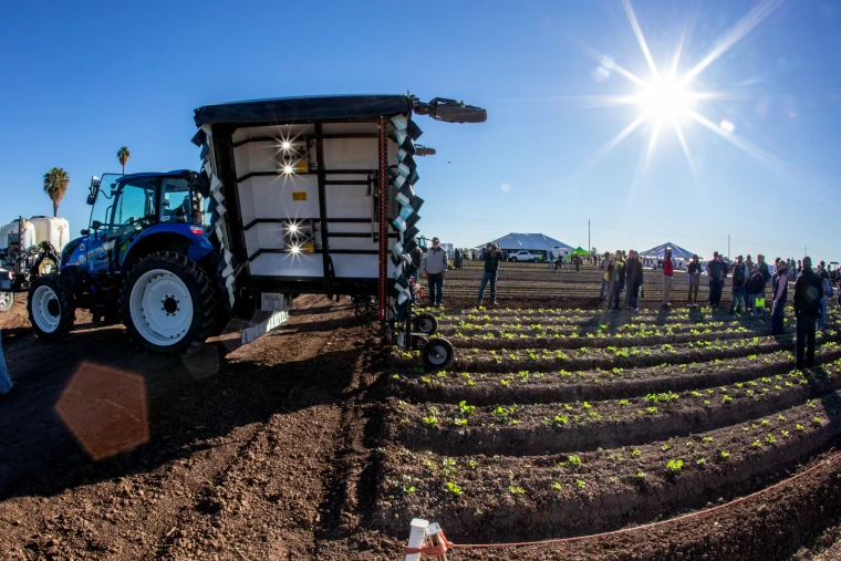 A large blue tractor is parked in a crop field as people observe farm machinery during a sunny outdoor event.