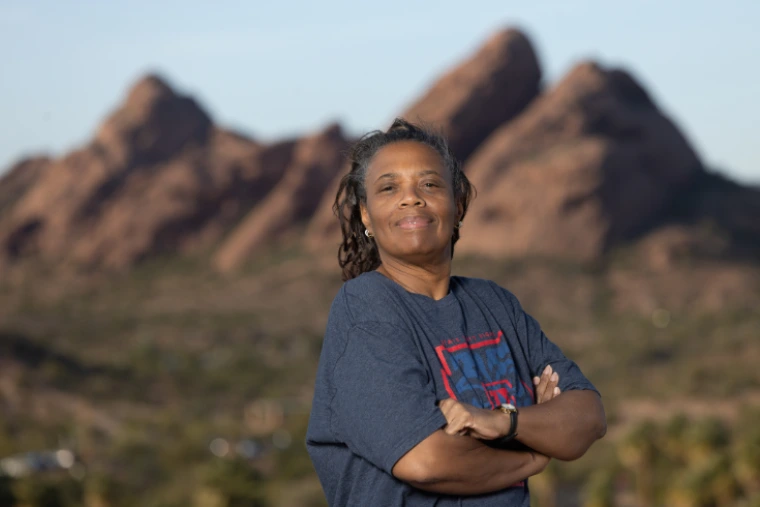 A person standing outdoors with arms crossed, wearing a casual T‑shirt, with large red rock formations and desert landscape visible in the background.