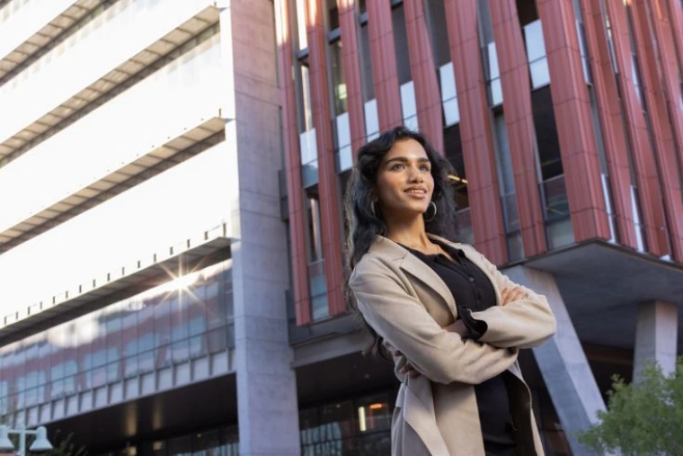 Gayatri Kaimal ’27 standing with arms crossed in front of a modern building with red vertical panels and concrete columns in daylight.