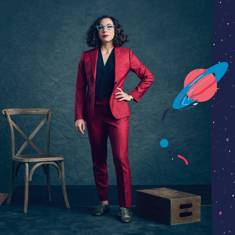 A person stands in a studio portrait wearing a red suit and blue-framed glasses against a dark backdrop. A wooden chair and a small platform sit beside the subject.