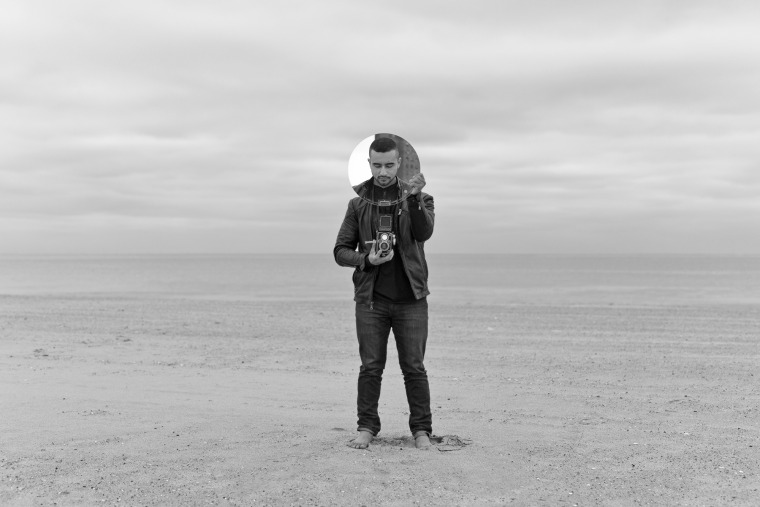 A photograph of poet, Javier Zamora, standing in the sand with a camera.