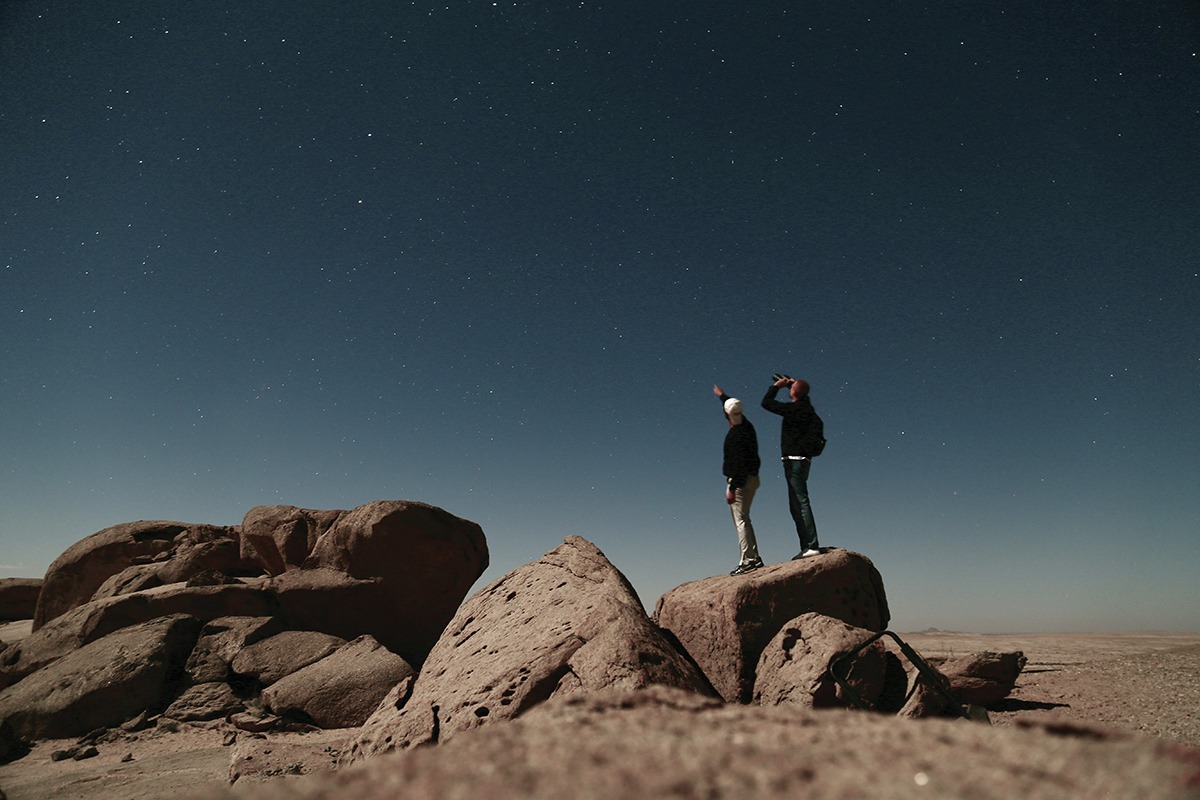 A photograph of two individuals standing on a large rock, pointing at the stars