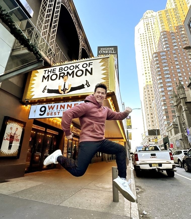 Photo of Alumni Tony Moreno ’21 jumping in front of a theater playing "The Book of Mormon" - the Broadway musical he is a "swing" on.
