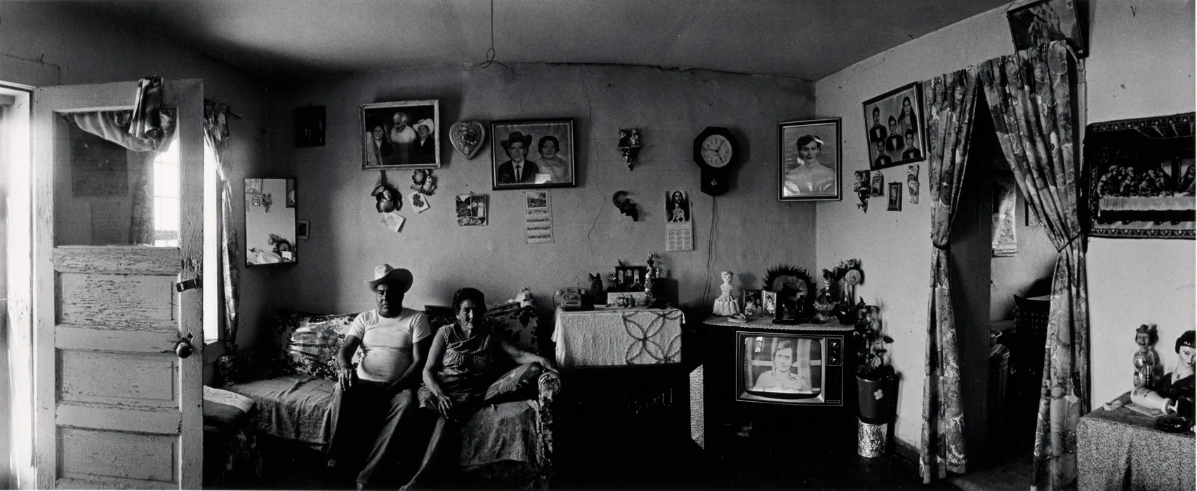 A photograph of a man and a woman sitting in a living room with family photos, a mirror, clock and television behind them.