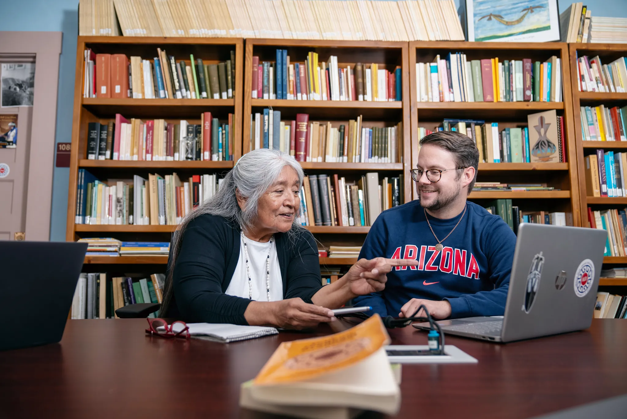 A woman talking and pointing to a computer while a man smiles at her in front of a shelf of books.