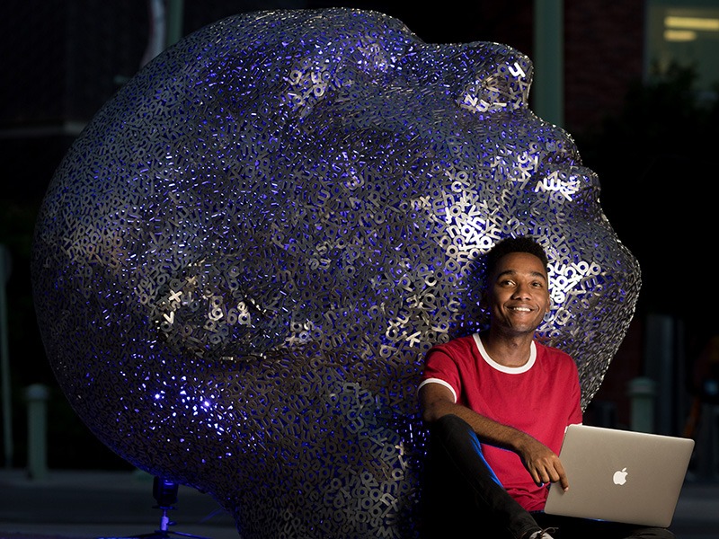 A photograph of Kameron Peyton looking up and smiling with a large human-size sculpture of a head behind him