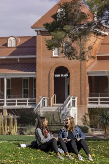 Two female students sitting on the grass in front of Old Main