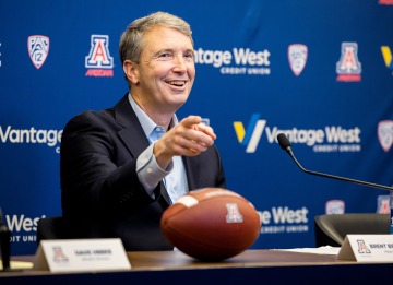 A man in a suit smiling and pointing with a football in front of him at a press conference 