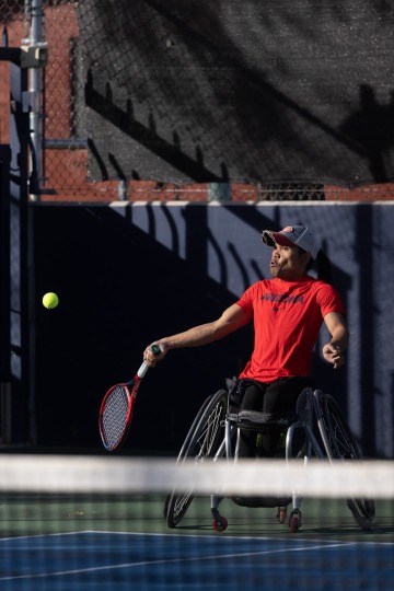 A photograph of a man in a wheelchair wearing a red shirt hitting a tennis ball passionately 