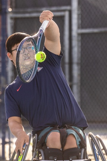 A photograph of a man in a wheelchair focused as he hits a tennis ball 