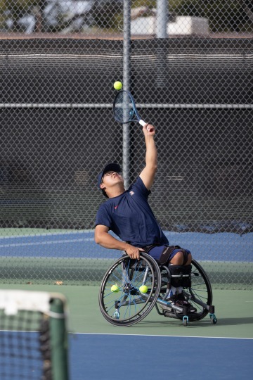 A photograph of a man in a wheelchair hitting a tennis ball above his head