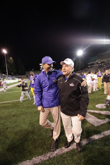 A man in a purple windbreaker side hugging another man in a black quarter zip on a football field at night 