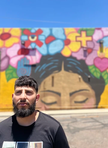 : A man stands in front of a colorful mural featuring closed eyes, flowers, and symbols on a sunny day.