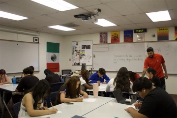 A teacher in a red shirt assists a student in a classroom filled with students working at tables. A Mexican flag hangs on the wall and a whiteboard displays the message, "Somewhere in the future your older self is looking at you to break the cycle.