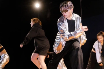 Dancer in a silver jacket and sunglasses performs under a spotlight, surrounded by fellow dancers in dynamic motion.