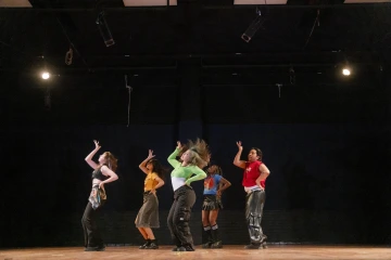 A group of six dancers strikes a synchronized pose on a dimly lit stage, each wearing vibrant, expressive outfits.