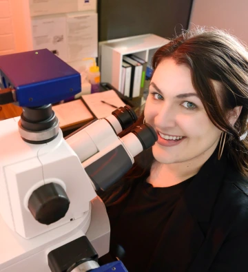 A smiling woman looks up from a high-powered microscope in a lab setting, with colorful cell imagery on the computer screen beside her.