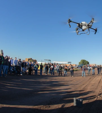 A crowd gathers to watch a flying agricultural drone demonstration on a farm, with clear skies and farm equipment in the background.