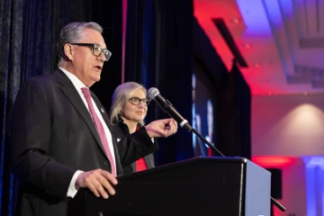 A man in a suit and red tie speaks at a podium with a microphone, while a woman stands behind him on stage. The background is lit in red, blue, and purple tones.