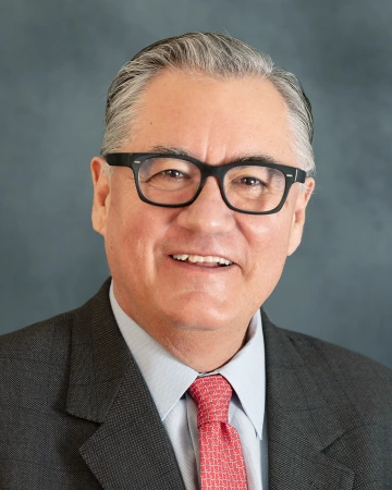 Professional headshot of a smiling man wearing glasses, a gray suit, and a red patterned tie against a blue background.