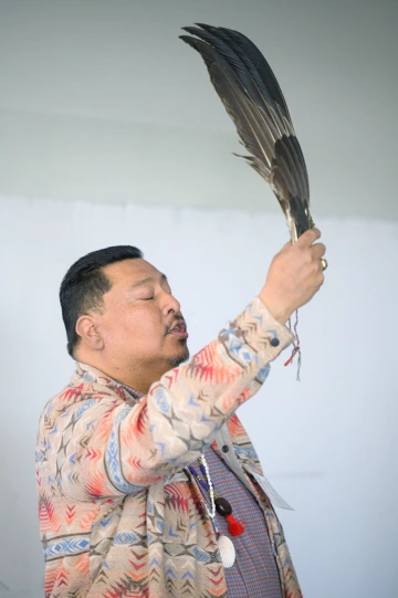 A person holding an eagle feather upright in a ceremonial gesture, wearing a patterned jacket and beaded necklaces, eyes closed in a moment of reflection.