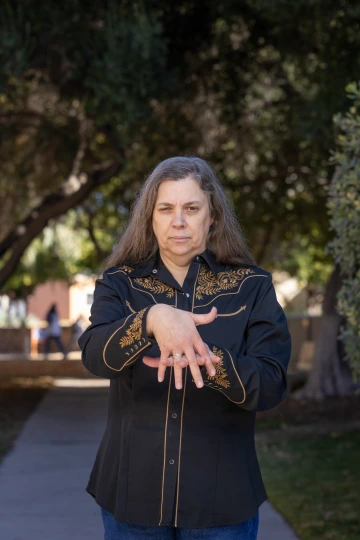 An unsmiling woman standing under a tree canopy giving the "Indigenous" sign in sign language. People are seen walking in the background.